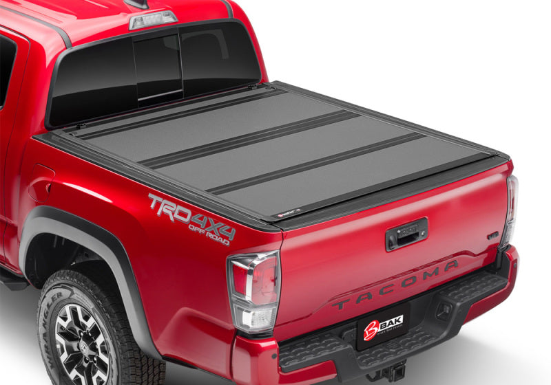 A sleek BAK 16-20 Toyota Tacoma 5ft Bed BAKFlip MX4 Matte Finish bed cover, featuring heavy-duty aluminum panels and a prominent BAK logo near the tailgate, enhances a red Toyota Tacoma pickup truck adorned with "TRD 4x4 Off Road" decals on the side. The visible rear wheels and taillights contribute to its rugged yet sophisticated appearance.