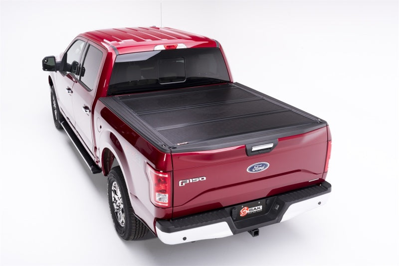 The image displays a red Ford F-150 truck from a rear angle set against a white background. This truck is equipped with the BAK 17-23 Ford Super Duty 6ft 9in Bed BAKFlip F1, a hard folding truck bed cover crafted from fiberglass reinforced polymer. The rims and side mirrors showcase black and chrome accents.