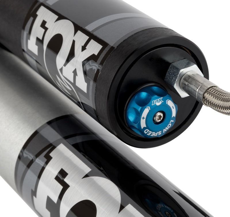 Close-up of the FOX 99+ Chevy HD 2.0 Performance Series rear shock absorber, ideal for enhancing off-road handling in trucks or SUVs, prominently displaying the FOX logo. One absorber showcases a blue adjustable low-speed knob and an attached silver braided cable. The sleek, polished metal finish enhances its robust design.