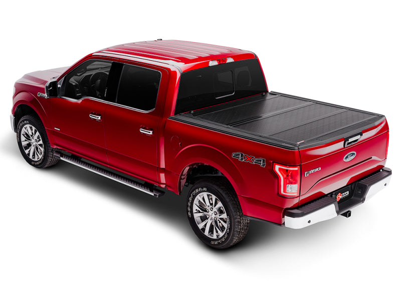 A red pickup truck equipped with a BAK 16-20 Nissan Titan 5ft 6in Bed BAKFlip G2 from BAK is seen from above and at an angle. The truck is parked on a white surface, highlighting its four doors and large wheels, providing an excellent cargo management solution.