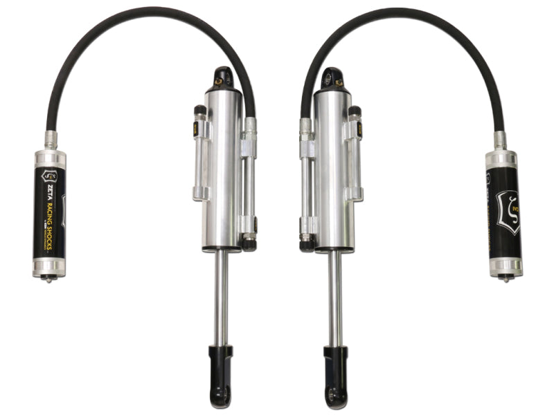 The image displays a pair of ICON 10-14 Ford Raptor Front Secondary 3.0 Zeta Series Shocks RR, showcasing silver, cylindrical automotive shock absorbers equipped with black flexible hoses. These shocks feature easy-to-adjust knobs and prominently display the ICON logo on their branded labels. They are positioned parallel against a plain white background, evoking the design elements of components used in the Ford SVT Raptor series.
