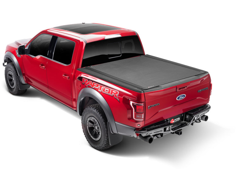 A red pickup truck with striking Raptor graphics on the side is shown. It features large black wheels, dual exhaust, and a sleek hard rolling cover over the bed. The BAK 12-18 Dodge Ram (19-21 Classic) w/ Ram Box Revolver X4s 6.4ft Bed Cover (2020 New Body Style) boasts matte black aluminum slats, viewed from a slightly elevated rear angle that showcases its robust build.