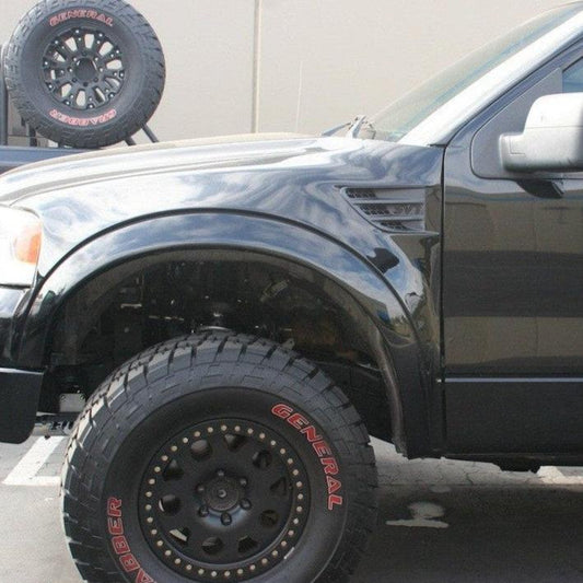 A close-up of the front right wheel and tire of a black pickup truck parked outdoors, showcasing its large, rugged General Grabber tire. The rear spare tire is visible mounted on a rack in the background. The focus is on the truck's suspension and ADV Fiberglass 2004-2008 FORD F-150 TO RAPTOR FENDERS from Advanced Fiberglass Concepts.