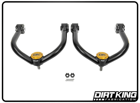 A set of 04-23 Nissan Titan Ball Joint Upper Control Arms in black, with gold caps, crafted by DIRT KING, is showcased on a white background. Positioned between them are two nuts and a cotter pin, which highlight the robust Moog ball joints. The DIRT KING logo appears in the bottom right corner.