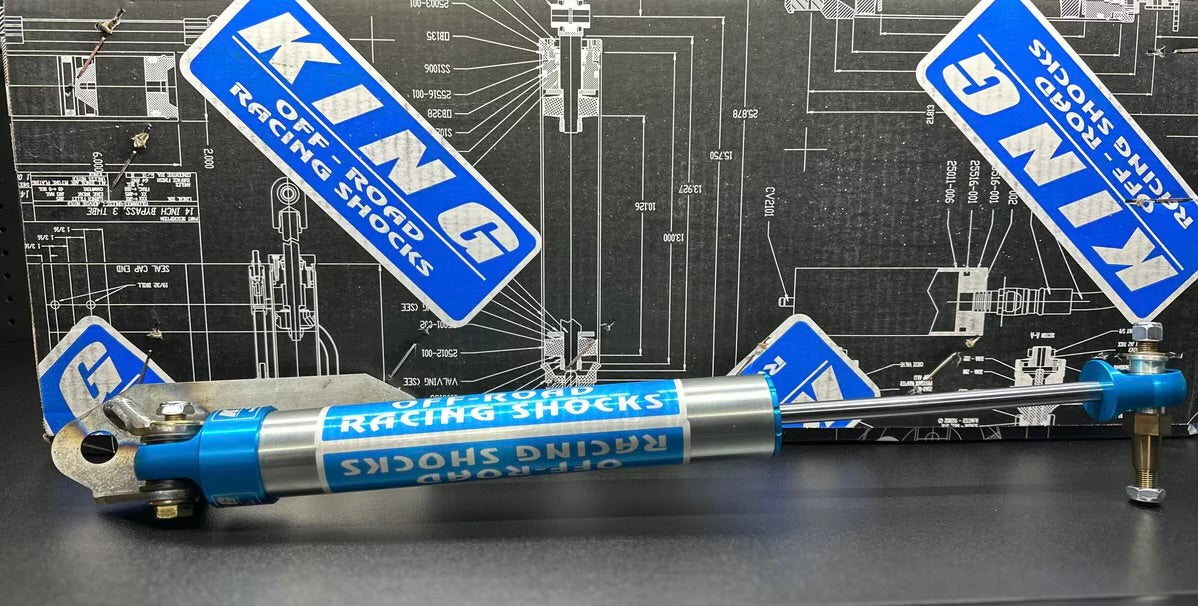 An LTW HD King Steering Stabilizer for 16-24 GM 2500/3500 HD models is displayed horizontally against a backdrop of technical blueprints and LTW Motorsports logo stickers. Featuring metallic and blue components, this stabilizer is engineered to enhance stability in rugged vehicle suspension.