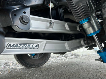 Close-up view of a vehicle's suspension system showcasing dual silver control arms with "Mazzulla Offroad" branding. The setup includes aftermarket extended travel shocks and the 2021+ RAM TRX Billet Trailing Arms, all part of an off-road vehicle, as indicated by the rugged terrain and background.