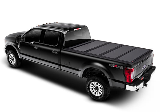 A black 4x4 pickup truck with a crew cab and long bed is equipped with the UV and scratch-resistant BAK 08-16 Ford Super Duty 6ft 9in Bed BAKFlip MX4 Matte Finish hard tonneau cover from BAK. Seen from a high angle, this vehicle, parked on a white background, highlights its rugged design and protective features.