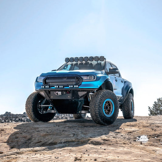 A robust, blue off-road truck equipped with large, rugged tires and multiple roof-mounted lights is parked on a rocky terrain under a bright sky. Featuring ADV Fiberglass 2019-2023 Ford Ranger Fenders and a modified front bumper, its lifted suspension emphasizes its capability for rough conditions.