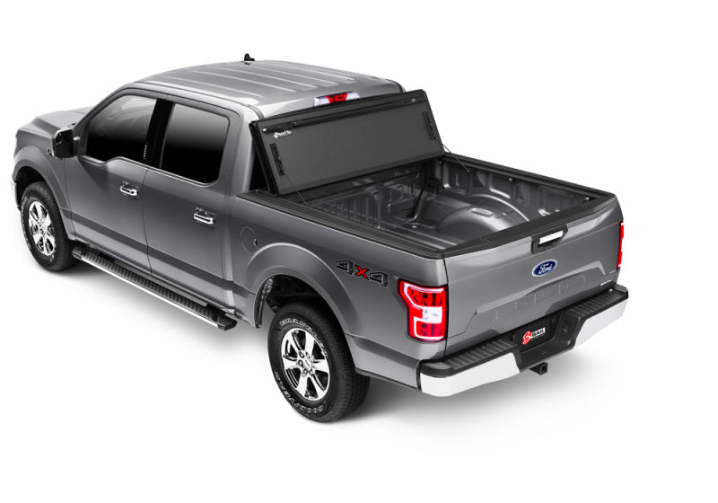 A rear-right angle view of a silver pickup truck with a crew cab showcases a 4x4 decal on its side. The BAK 15-20 Ford F-150 5ft 7in Bed BAKFlip MX4 Matte Finish truck bed cover by BAK, featuring sturdy aluminum matte finish panels, is partially opened. The tailgate is lowered, exposing the ample truck bed.