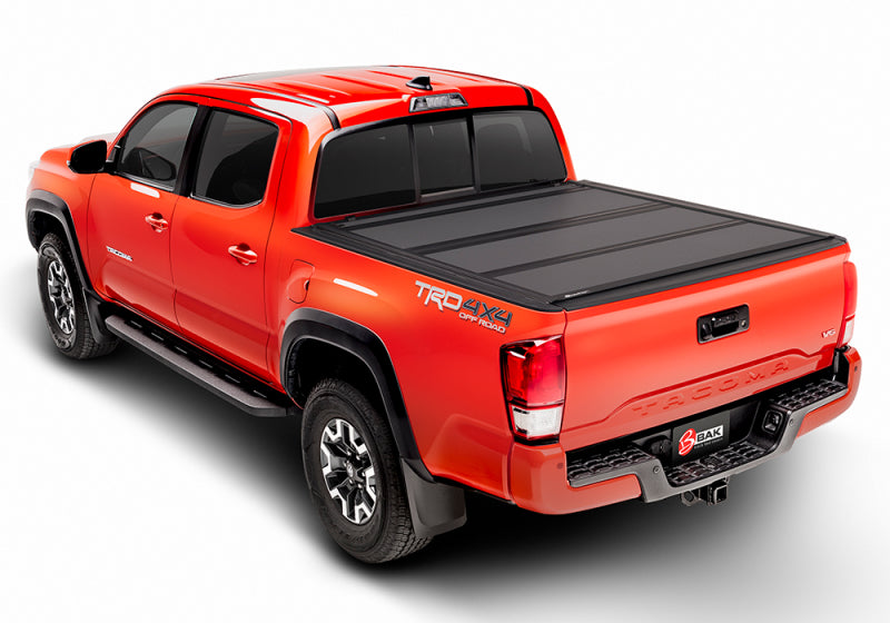 A red pickup truck featuring a BAK 05-15 Toyota Tacoma 5ft Bed (w/o Universal Tailgate Function) BAKFlip MX4 Matte Finish tonneau cover made of heavy-duty aluminum is adorned with a 4x4 decal on the side. Positioned at an angle, it emphasizes its rear and side profile with off-road tires and black side steps, highlighting its rugged versatility.