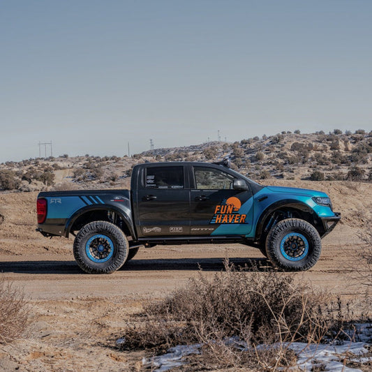 A modified black and blue pickup truck with rugged tires, aftermarket body panels, and a "FUN HAVER" decal on the side is parked on a dirt road in a desert landscape. The truck features off-road accessories, fiberglass fenders from ADV Fiberglass, and 2019-2023 FORD RANGER BEDSIDES amidst sparse vegetation and distant hills.