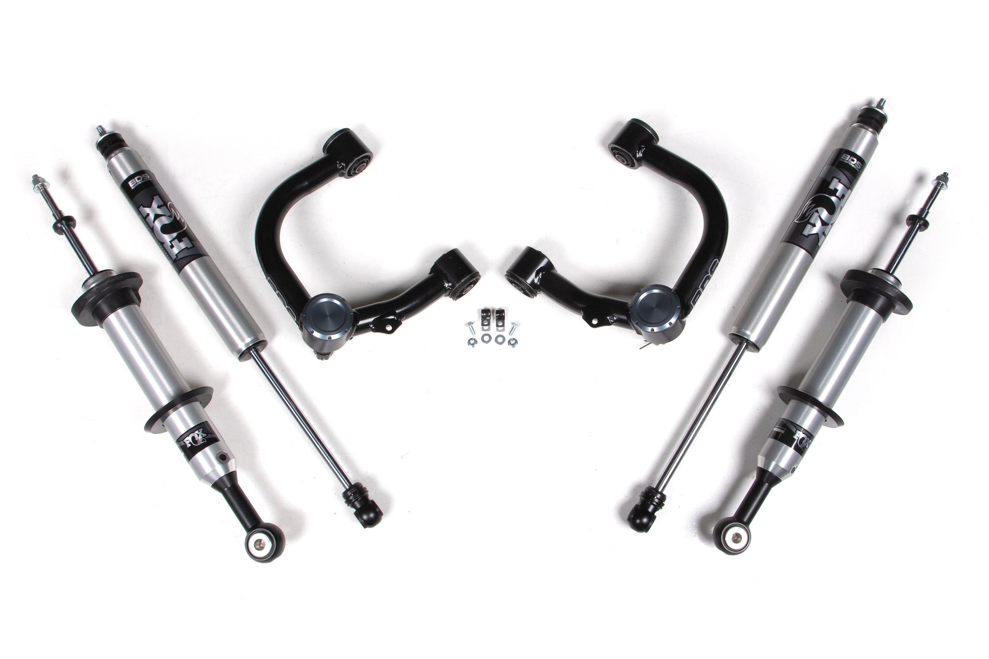 The image showcases a set of automotive suspension components laid out symmetrically on a white background. The components include four FOX 2.0 Performance shocks, two upper control arms, and a group of smaller hardware pieces such as bolts and nuts, all ideal for the BDS 1 Inch Lift Kit | FOX 2.0 Snap Ring Strut designed for Toyota Tacoma (2016-2023) 4WD models.