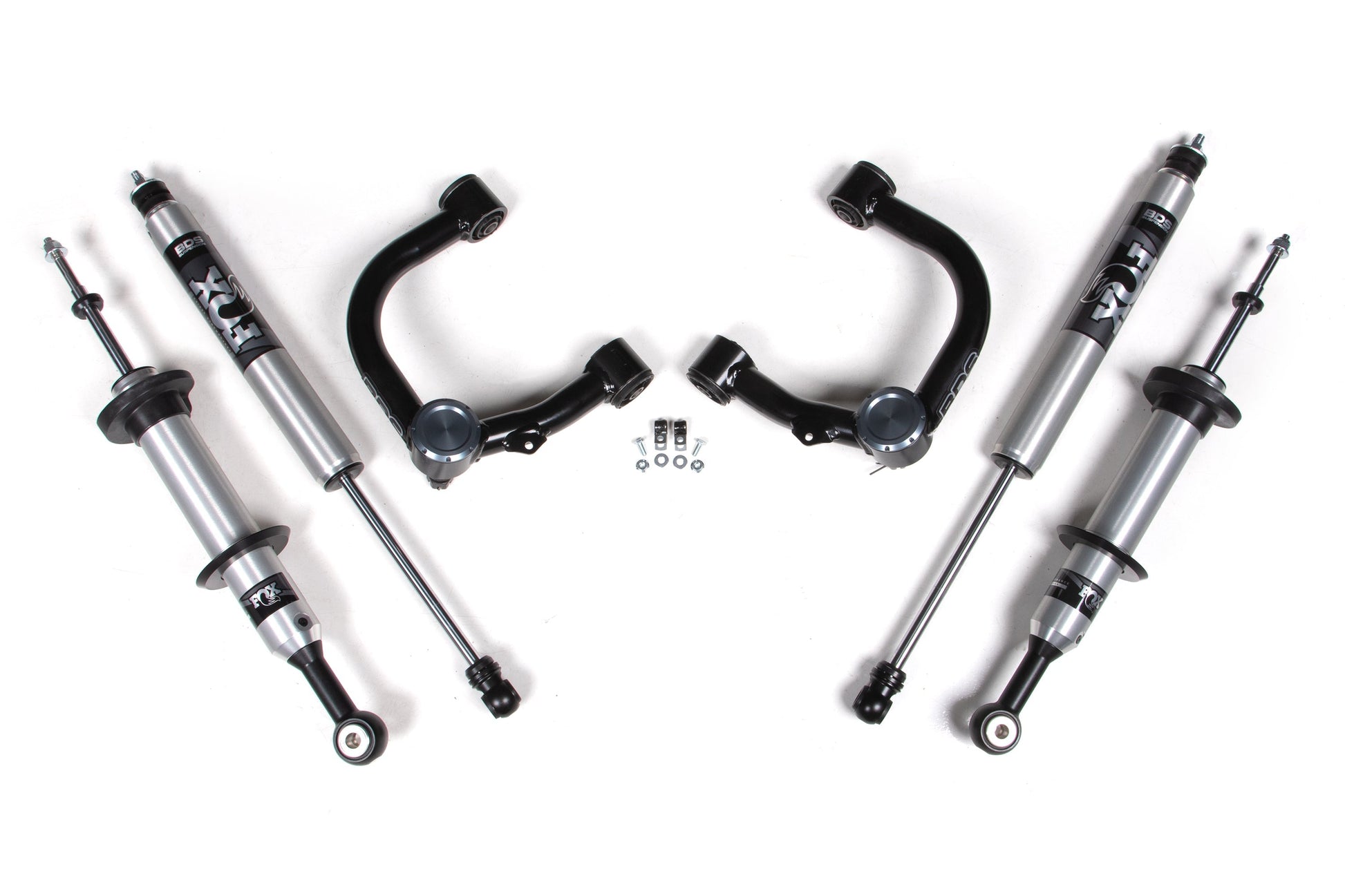 The image showcases a set of vehicle shock absorbers and suspension components, including four silver and black FOX 2.0 Performance shocks and two black control arms from the BDS 2 Inch Lift Kit with FOX 2.0 Snap Ring Strut for Toyota Tacoma (05-15) 4WD. Small bags with nuts, washers, and bolts are neatly arranged in the center, all displayed on a white background.