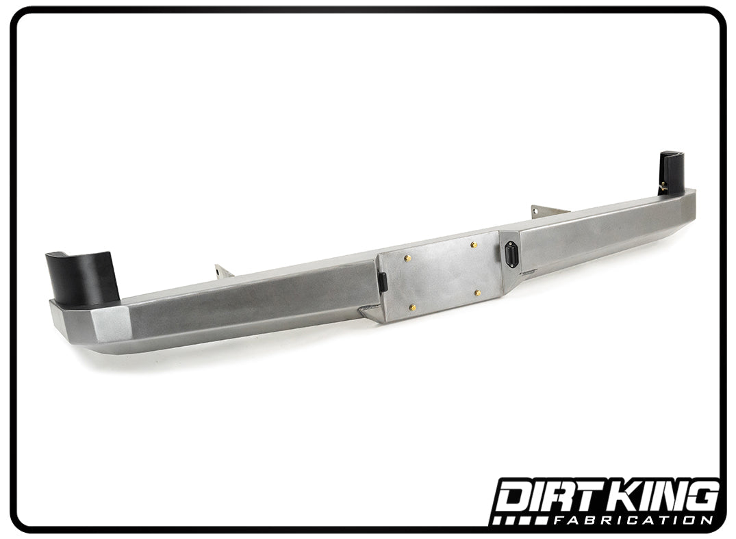 A DIRT KING 05-23 Toyota Tacoma Plate Rear Bumper is pictured with a black logo on the bottom right corner reading "Dirt King Fabrication." The bumper features a sleek, angular design that provides increased ground clearance, with mounting points on both ends and a central section designated for a license plate and accessories.