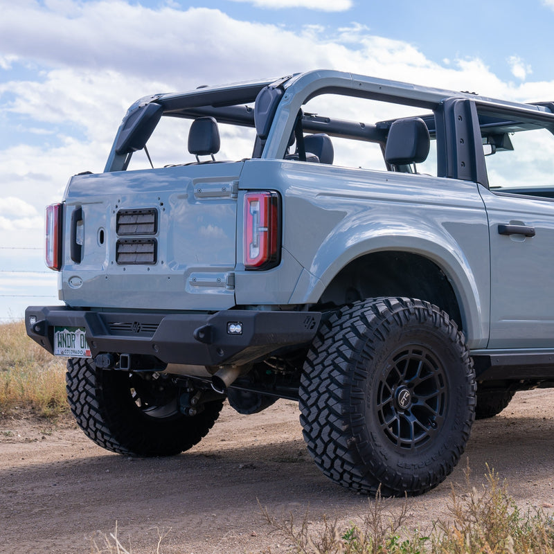 A light gray off-road SUV with an ADV Fiberglass 2021-2023 Ford Bronco 2-Door Rear Quarter Kit and the top and doors removed is parked in a dirt area. The vehicle has large, rugged tires and is equipped with a rear tow hook. The sky is partly cloudy in the background.