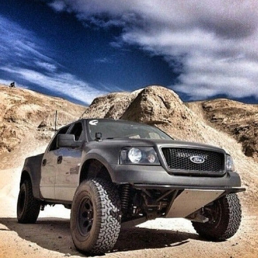 An ADV Fiberglass 2004-2008 FORD F-150 equipped with prerunner fender flares and off-road tires is parked in a desert landscape, set against sand dunes and a cloudy sky. The vehicle's dark paint contrasts starkly with the light, sandy terrain.