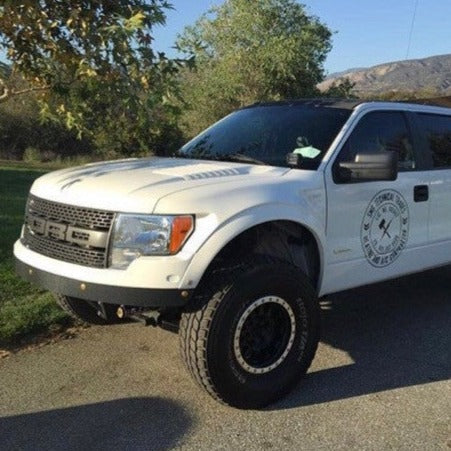 A 2010-2016 Ford Raptor, featuring a fiberglass hood by ADV Fiberglass and large tires, is parked on a paved road. The truck's door displays a logo and text, with trees and hills visible in the background under a clear blue sky.