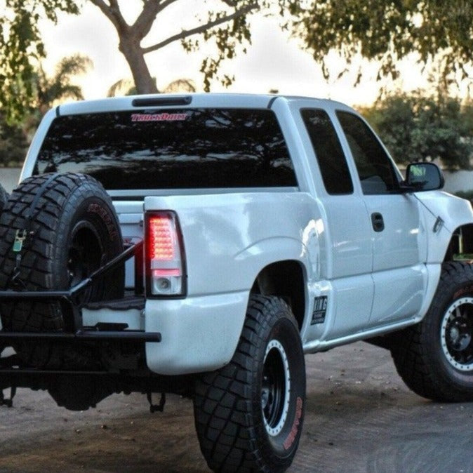 A lifted white pickup truck, outfitted with ADV Fiberglass 1999-2006 Chevrolet Silverado bedsides, is parked outdoors. It features rear off-road tires mounted on the bed, dark-tinted windows, and modifications for off-roading such as large tires and a custom bumper. Trees and greenery create a serene backdrop.