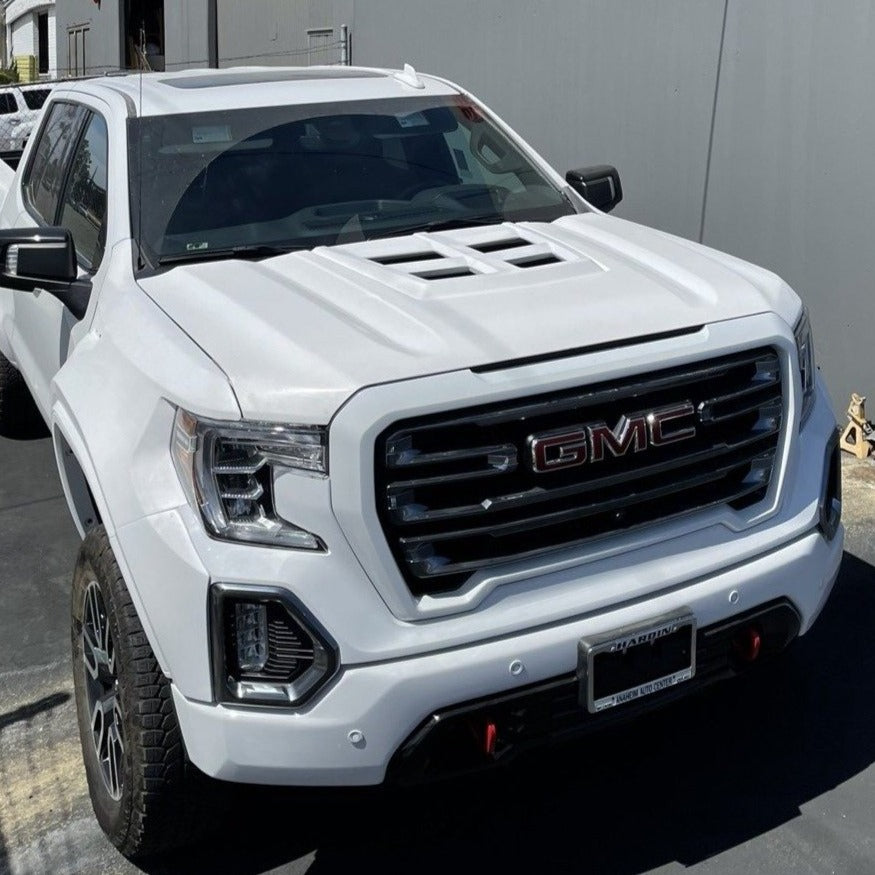 A white GMC pickup truck is parked in front of a gray building. The vehicle boasts a prominent front grille with the GMC logo, black accents around the headlights, and rugged tires. Notably, it features an ADV Fiberglass 2019-2021 GMC Sierra Hood with air vents and red tow hooks on the front bumper.