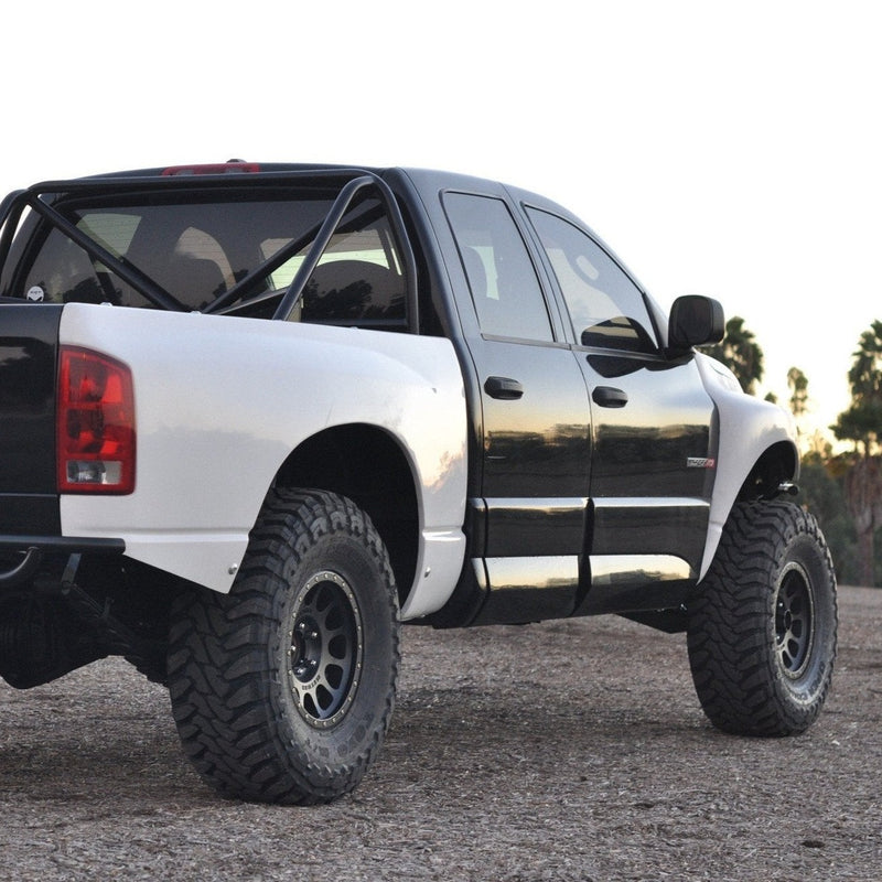 A white and black off-road truck with large, rugged tires and a lifted suspension is parked on a gravel surface, sporting ADV Fiberglass 2003-2008 Dodge Ram bedsides. The truck has a roll cage visible in the bed area, and a clear sky is seen in the background.