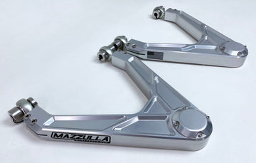 A pair of silver aluminum upper control arms are arranged on a white surface. The arms have a sleek design with angular lines and multiple points of adjustment. Featuring uniball pivots, they are part of the 2007-2018 CHEVY/GMC 1500 LONG TRAVEL kit for offroad vehicles. One arm has "MAZZULLA OFFROAD" branded on it.