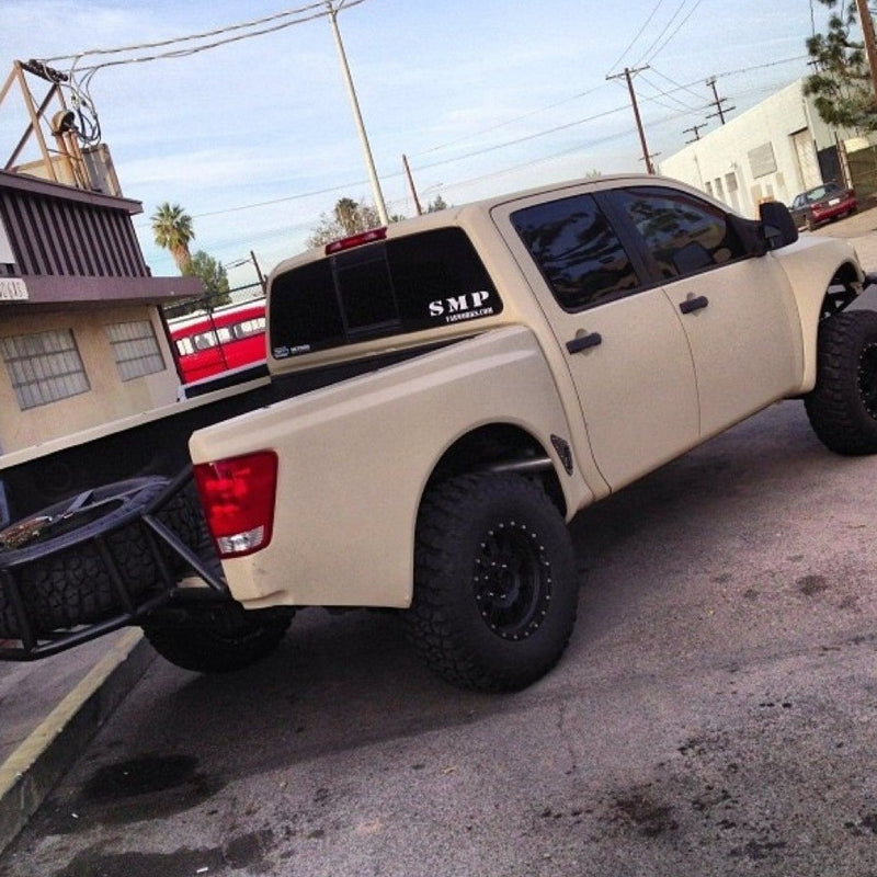 A beige, lifted Nissan Titan pickup truck with large off-road tires is parked in front of a building. The truck bed, equipped with ADV Fiberglass 2004-2014 NISSAN TITAN BEDSIDES-prerunner bedsides, has a mounted black spare tire. The back window features a decal reading "SMP." The sky is clear and the location appears to be an industrial area.