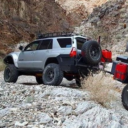 A 2003-2009 TOYOTA 4-RUNNER with ADV Fiberglass Fenders, featuring large tires and a roof rack, navigates a rocky terrain. It is towing a trailer loaded with supplies, indicating an off-road adventure or excursion. The surrounding landscape features rocky hills and sparse vegetation.