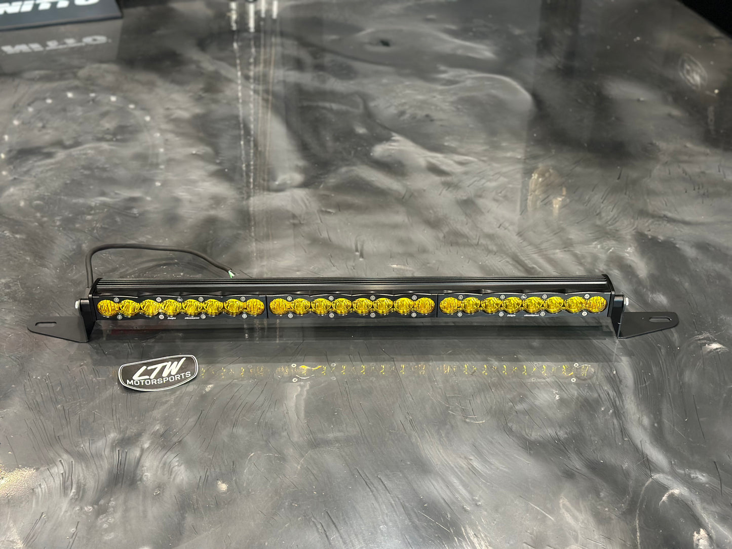 A 30-inch Baja Designs LED light bar, featuring multiple yellow lenses, is mounted on a dark, reflective surface using the LTW Motorsports Behind the Grille Light Bar Mount Kit designed for the 22-24 Chevy Silverado 1500. The "LTW Motorsports" logo is prominently displayed near the light bar, enhancing its sleek presence against the blurred background and evoking a Chevy Silverado 1500 setup.