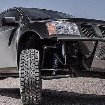 Close-up view of a rugged off-road vehicle showcasing its raised suspension and large all-terrain tire. The vehicle boasts ADV Fiberglass 2004-2014 NISSAN TITAN FENDERS, an aftermarket black metal bumper, visible shock absorbers, and a sleek, sturdy exterior. The shot focuses on the front left side, highlighting its durable build.