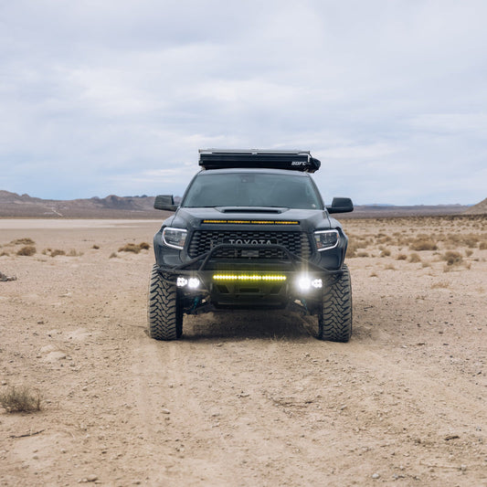 A black Toyota 4Runner, modified for off-road adventures with large tires and extra lighting, is parked on a dirt road in a barren desert landscape. The vehicle features ADV Fiberglass fenders designed for the 2014-2021 Toyota Tundra and includes a roof rack. Distant mountains are visible under the cloudy sky in the background.