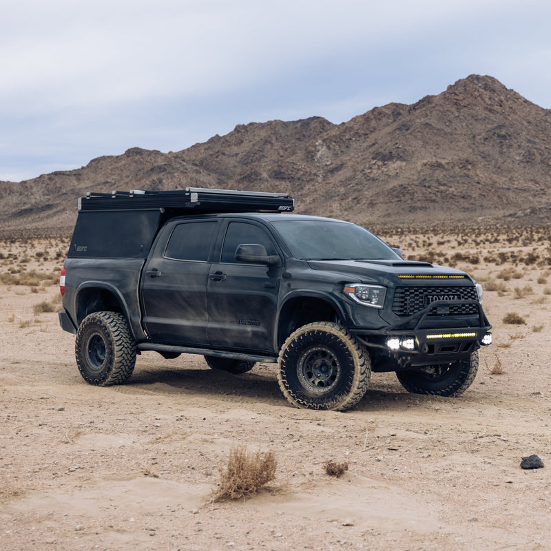 A black off-road truck with a camper shell and large all-terrain tires is parked on a desert terrain with rocky mountains in the background. Outfitted with parts for a 2014-2021 Toyota Tundra, including ADV Fiberglass aftermarket bedsides, LED lights, and roof rails, it showcases a rugged, adventurous setup.