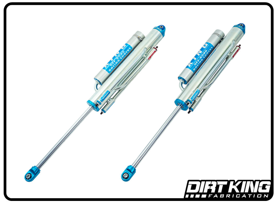 Image of two Dirt King Bed Cage Spec King 16" Triple Bypasses, featuring a blue and silver color scheme with remote reservoirs. Designed by DIRT KING SHOCKS for off-road use, these bypass shocks enhance ride quality. The Dirt King Fabrication logo is visible in the bottom right corner of the image.