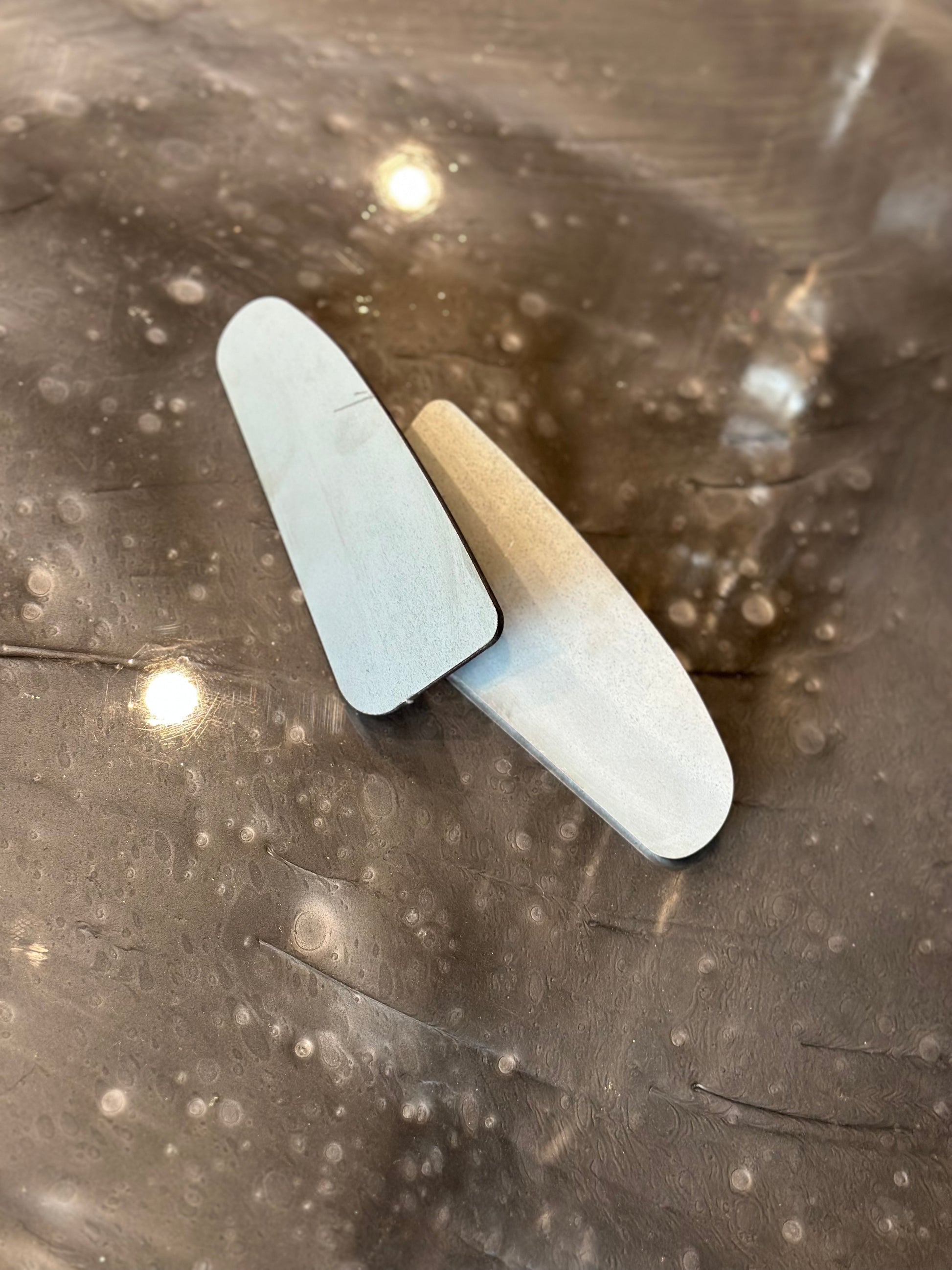 Two wooden wedges are positioned on a reflective, speckled surface. The wedges, reminiscent of the precision found in LTW Motorsports' 05-22 Tacoma Body Mount Chop Plates (Weld On), are pale in color and have a smooth texture. The surface they rest on has visible marks and small circular reflections, possibly from lighting above.