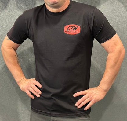 A person stands confidently with hands on hips, showcasing the LTW Black w/ Red Logo Shirt by LTW Motorsports, featuring a striking red logo on the front, set against a plain, textured wall.