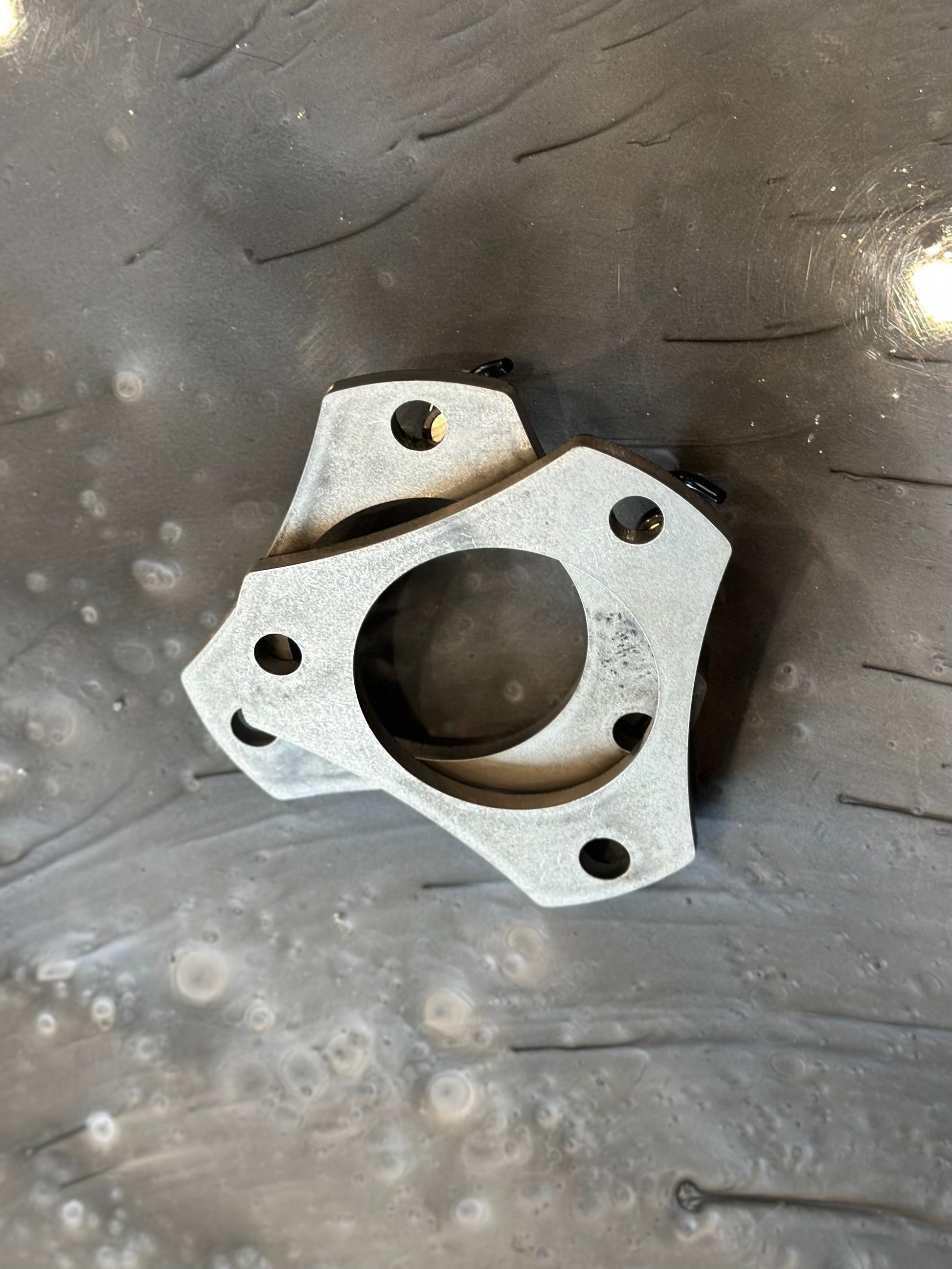 A 05-18 Tacoma 1/4" Top Hat Spacer (Pair) by LTW Motorsports, featuring a multiple-hole design that resembles a mounting bracket, lies on a dark, textured surface. The component includes five edges and a central circular opening, making it suitable for models like the Tacoma from 2005 to 2018.