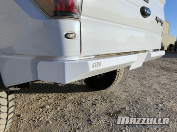 A white Ford Raptor truck is parked on a gravel surface in front of a beige brick wall. The truck boasts a sleek design with large off-road tires, a CNC-cut steel bumper for increased approach angle, and tinted rear windows. "MAZZULLA OFFROAD" branding is visible in the top left corner of the image, indicating its premium 2009-2014 FORD F-150/RAPTOR REAR PLATE BUMPER.