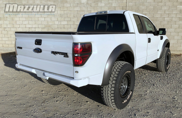 A white Ford Raptor truck is parked on a gravel surface in front of a beige brick wall. The truck boasts a sleek design with large off-road tires, a CNC-cut steel bumper for increased approach angle, and tinted rear windows. "MAZZULLA OFFROAD" branding is visible in the top left corner of the image, indicating its premium 2009-2014 FORD F-150/RAPTOR REAR PLATE BUMPER.