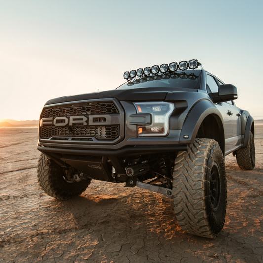 A rugged black truck equipped with ADV Fiberglass's 2017-2020 Ford Raptor Fenders and lifted suspension stands boldly on the dry, cracked desert surface. The sunset casts a warm glow, highlighting the vehicle's aggressive front grille and off-road tires.