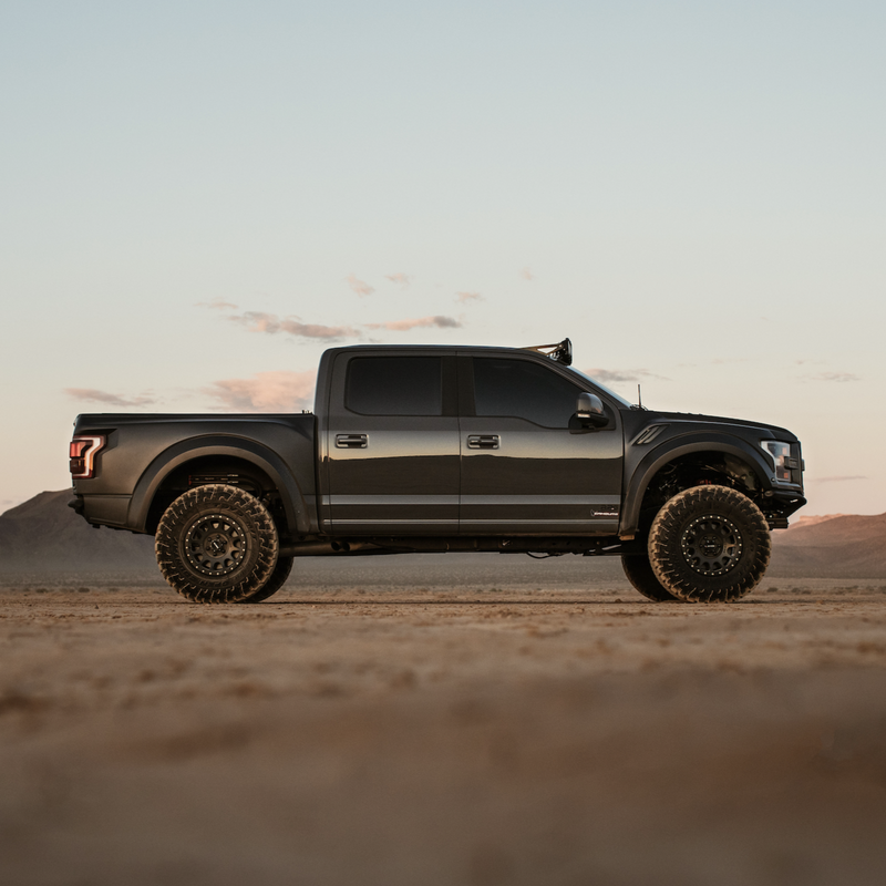 A Ford Raptor from the 2017-2020 model range, featuring ADV Fiberglass bedsides and large off-road tires, is parked on a sandy terrain under a clear sky, with low mountains in the background. The truck faces left, highlighted by soft natural light.