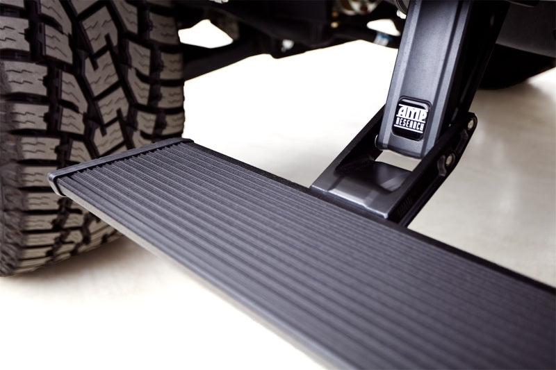 A close-up of a Chevrolet Silverado featuring the AMP Research 2019 Chevy Silverado 1500 Crew PowerStep Xtreme in black with OEM-style illumination, and large, textured tires. The PowerStep Xtreme showcases a ribbed surface for enhanced grip and prominently displays the AMP Research logo against the smooth, light-colored surroundings.