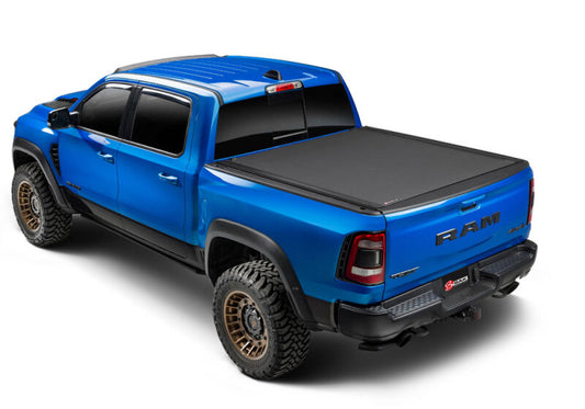 A blue RAM pickup truck, equipped with the BAK 14-18 GM Silverado/Sierra Revolver X4ts tonneau cover, features large off-road tires and a sleek design. Viewed from the rear at a slightly elevated angle, the black cover enhances its polished appearance, demonstrating an impeccable blend of style and cargo management for rugged terrain.