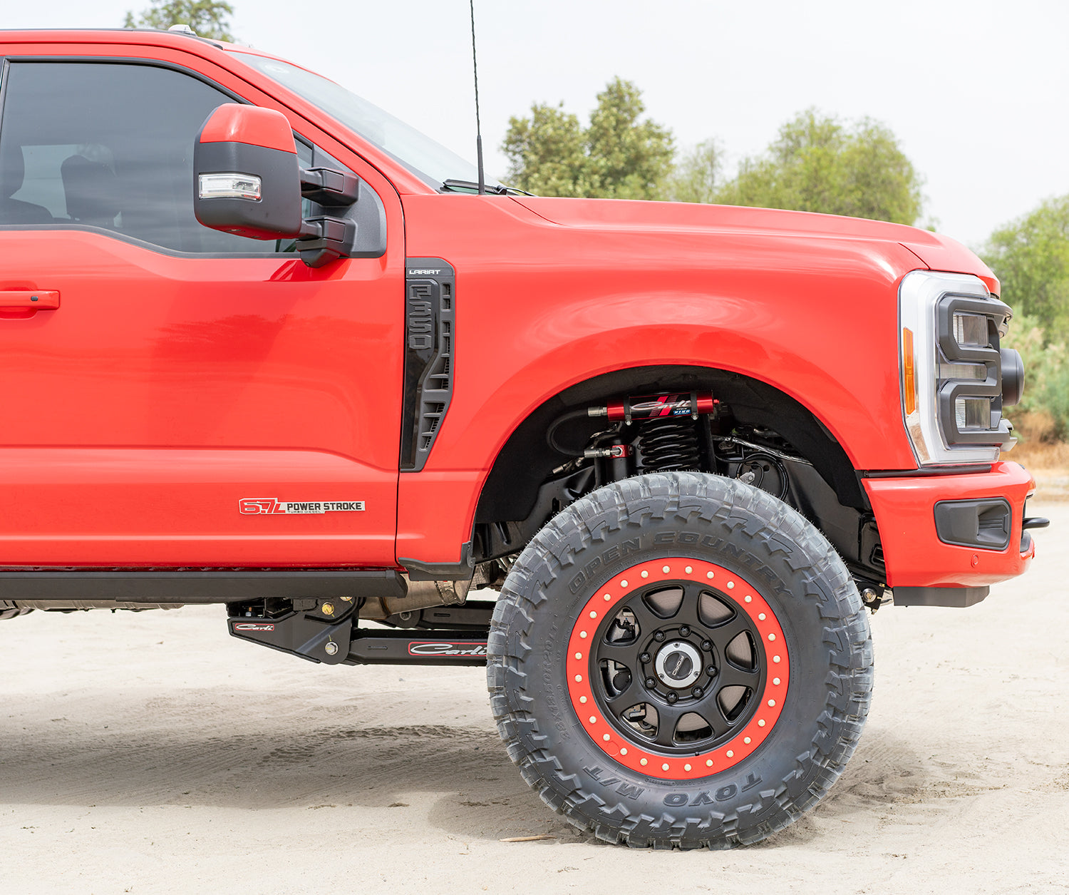 A close-up of a red truck with large off-road tires and black rims, parked on a sandy surface, showcases its impressive features. The vehicle is equipped with Carli Suspension's 5.5" Lift and Radius Arm Drop Brackets for the 23+ Ford F-250/F-350 (4WD), proudly displaying the "Power Stroke" logo. The backdrop of trees under a cloudy sky emphasizes the powerful presence of this truck.