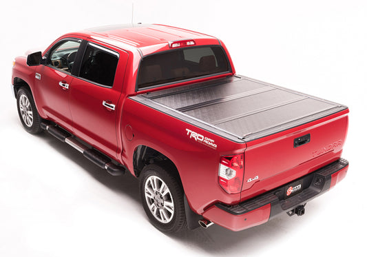 A red pickup truck, featuring a BAK 05-15 Toyota Tacoma 5ft Bed BAKFlip G2 from BAK, is displayed from an elevated rear angle. It sports a TRD Off-Road logo on the side, chrome wheels, and side steps. The truck is fitted with a sturdy bed cover to ensure maximum theft protection and is parked against a plain white background.