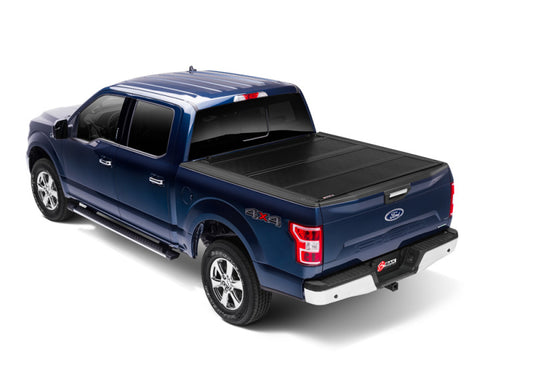 A blue Ford 4x4 pickup truck with a four-door cabin and a covered cargo bed is displayed from a rear three-quarter angle. The tailgate features the iconic Ford emblem, and its sturdy design is complemented by the BAKFlip FiberMax aluminum hard folding truck bed cover by BAK, designed for the 15-20 Ford F-150 with a 5ft 6in bed.