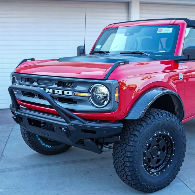 A bright red Ford Bronco with customized off-road features, including an ADV Fiberglass 2021-2023 Ford Bronco heat extractor hood, is parked in front of a garage with white doors. The vehicle also boasts a roll bar, elevated bumper, textured black fender flares, large off-road tires, and a black hood decal.