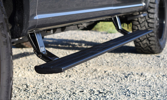 The image highlights a black truck equipped with an AMP Research Smart Series PowerStep, designed for the 19-23 Ford Ranger and 21-23 Ford Bronco (excluding Raptor models). The electric-powered running board features a retractable side step made of metal and plastic, positioned beneath the door on a gravel surface. The large tire is visible to the left of the step.