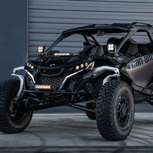 A Can-Am Maverick with large tires and a rugged black exterior is parked by a metal garage door, equipped with the Baja Designs Can-Am Maverick R XL Pro A-Pillar Kit. It features multiple LED lights and an aggressive design, ideal for tackling adventurous terrain.