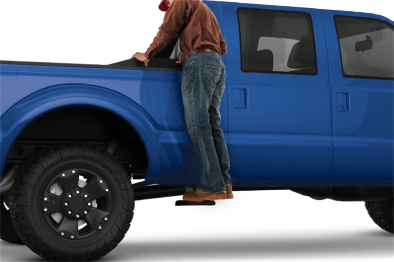 A person wearing a red cap, brown shirt, blue jeans, and brown boots is climbing into the bed of a blue pickup truck. The truck features large black tires and an AMP Research 19-22 Ram 1500 (Excl. TRX) BedStep2 in black aiding their ascent.