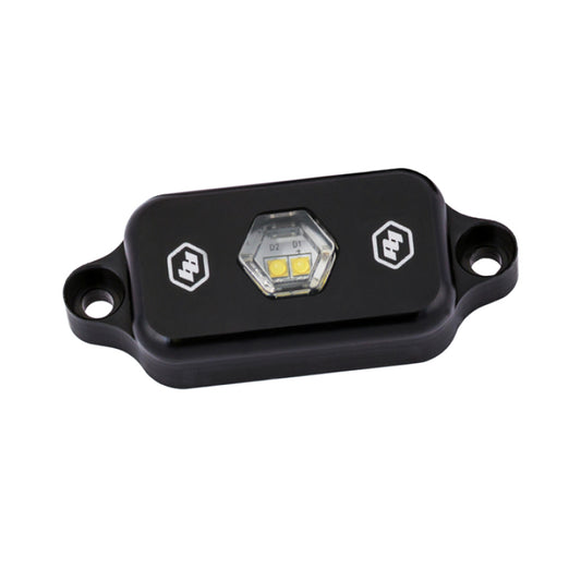 The Baja Designs LED Rock Light - Clear is a compact, black device with a hexagonal clear cover that showcases two LED bulbs. It features dual mounting holes for easy installation and prominently displays two logos on its surface.