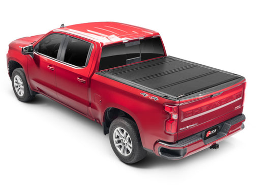 A red Chevy Silverado with a four-door cab is shown from a rear angle. It sports silver rims and a black BAK BAKFlip G2 hard folding truck bed cover for full bed access. The tailgate displays brand markings and features 4x4 decals. For added peace of mind, the BAKFlip G2 cover provides maximum theft protection.